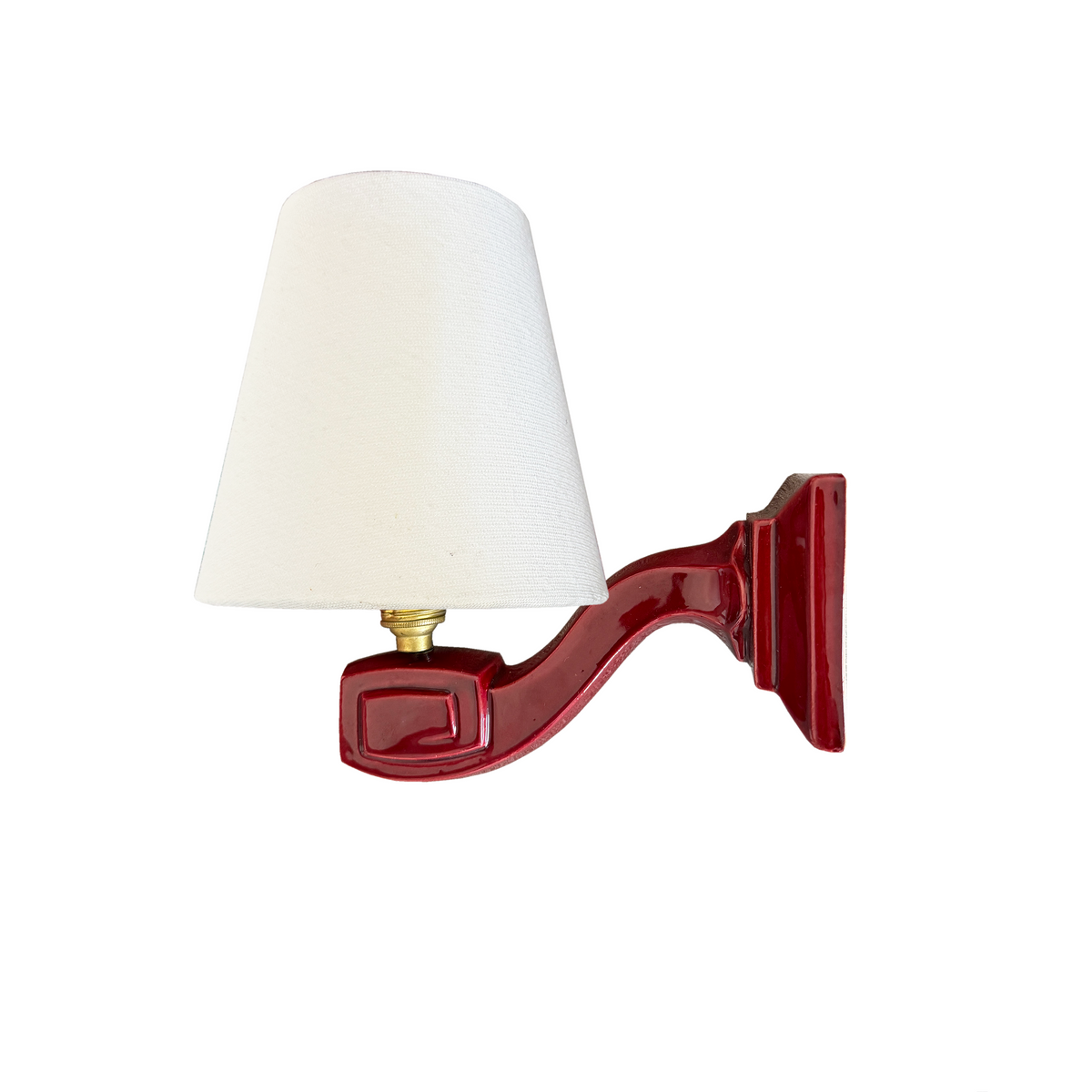 A wall-mounted sconce with a tapered off-white fabric shade and a glossy deep red ceramic bracket, featuring a small gold-toned metal connector.