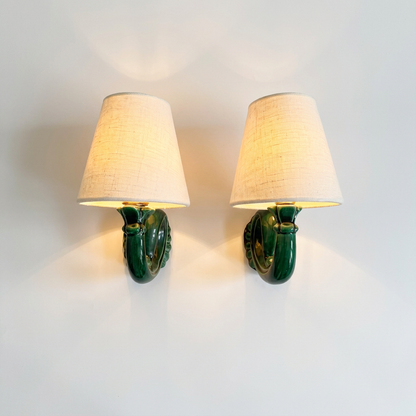 A pair of green ceramic wall sconces with tapered beige fabric shades, shown lit against a plain white wall.