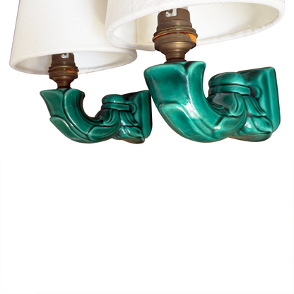 A close-up of a pair of vintage-style wall sconces with glossy teal green ceramic scrollwork bases and white fabric drum shades, shown against a plain white background.